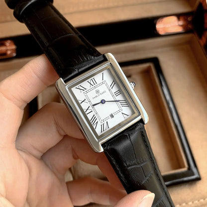 Rectangular watch with black leather strap held by a hand, with a brown box in the background.