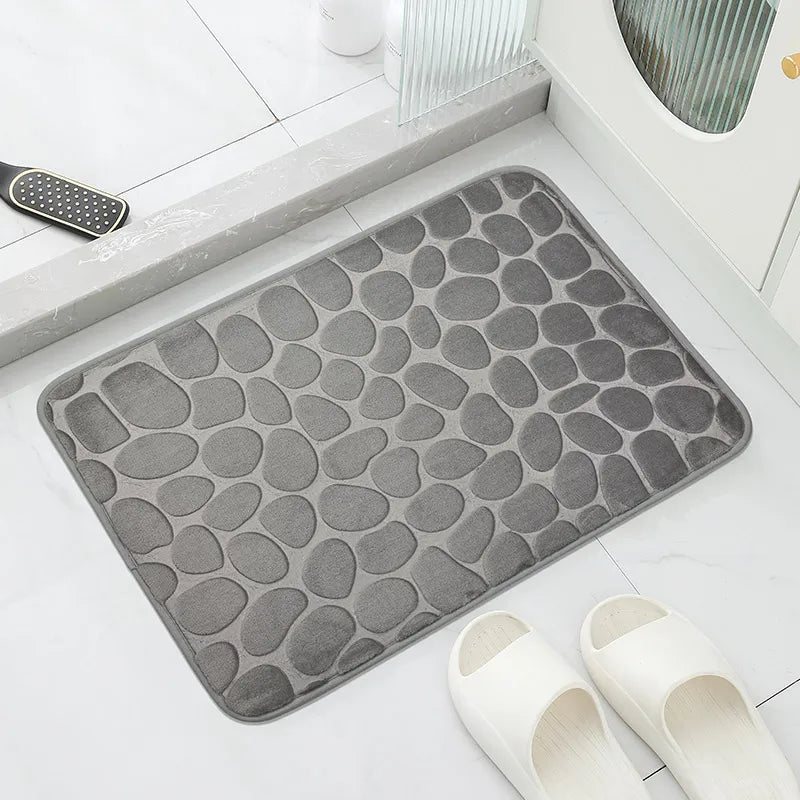 Gray textured bath mat on a bathroom floor with white slippers nearby.