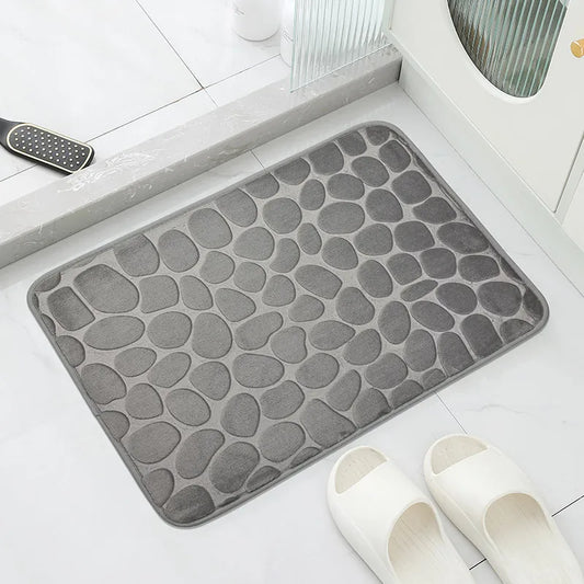Gray textured bath mat on a bathroom floor with white slippers nearby.