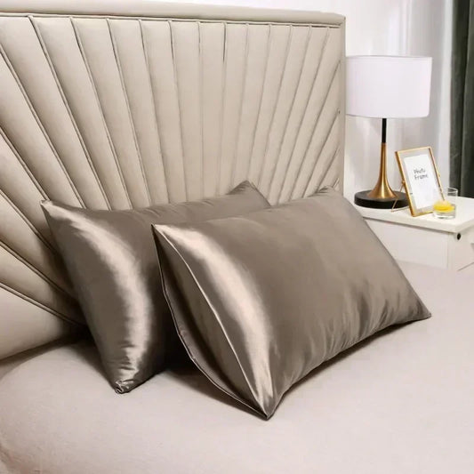 Two satin pillows on a bed with a lamp and decor items in the background.