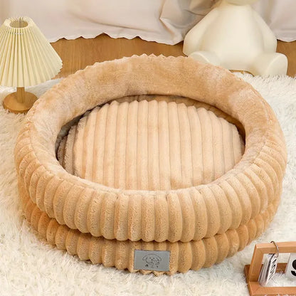 Beige pet bed on a fluffy white surface with a lamp and books in the background.