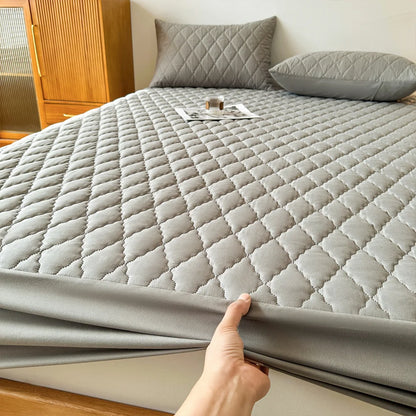 Person holding a quilted mattress pad over a bed with gray bedding.