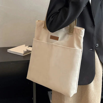 Beige tote bag held by a person wearing a black coat and beige pants.